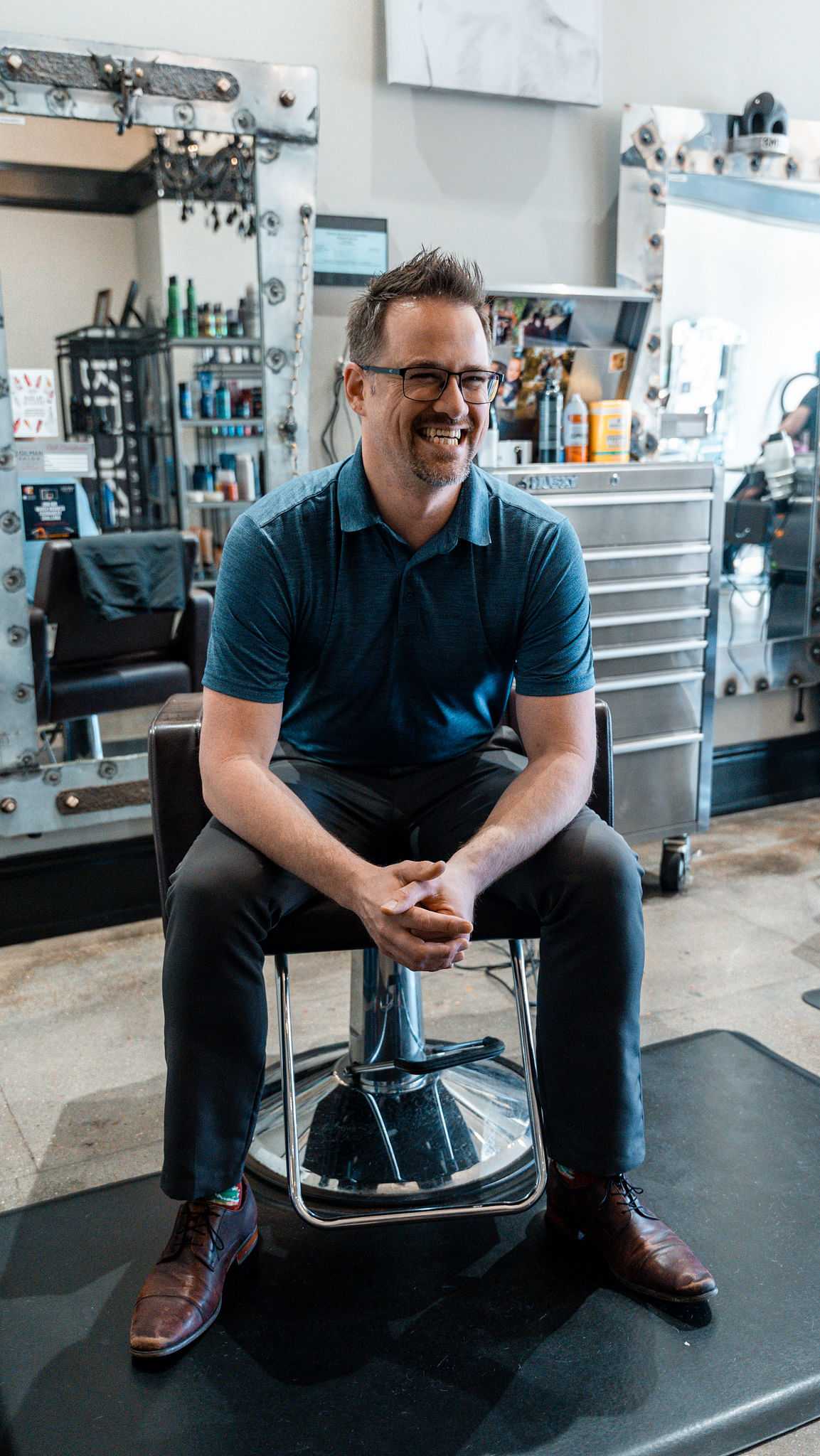Man sitting in a salon chair, smiling, with styling products and mirrors in the background.